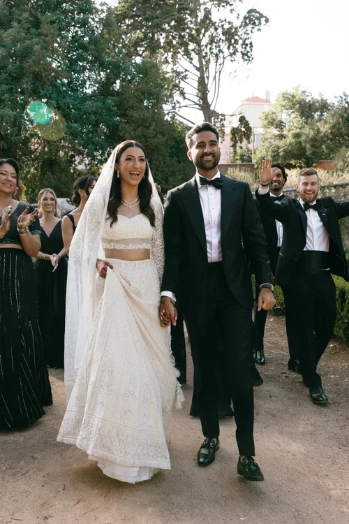 Bride in white embroidered dress and groom in black tuxedo walking through tropical gardens with wedding guests in Marbella
