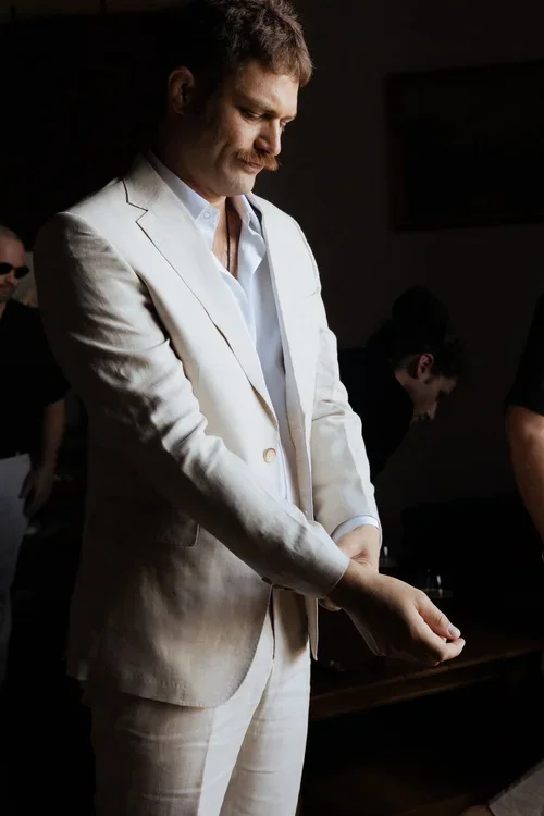 Groom in beige linen suit adjusting his cuffs in a moody wood-panelled library while getting ready on his wedding morning