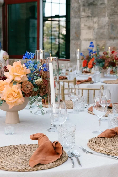 Elegant Marbella wedding reception table with peach roses, blue delphinium, orange poppies, terracotta linen napkins, woven placemats and crystal glassware at a stone venue