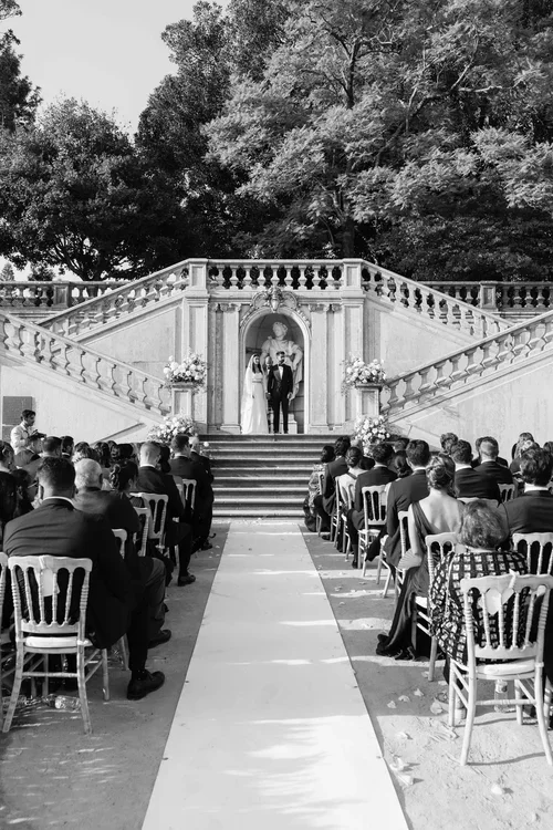 Outdoor wedding ceremony with bride and groom standing before a grand double stone staircase and marble statue niche flanked by white floral arrangements with guests seated on either side of a white aisle