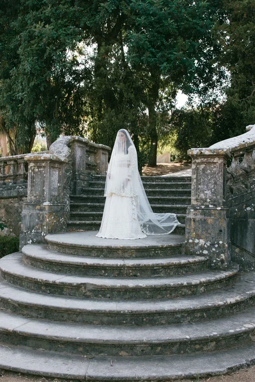 Bride standing on ancient stone steps in a long embroidered cathedral veil surrounded by cypress trees, editorial bridal portrait photography Andalusia Spain