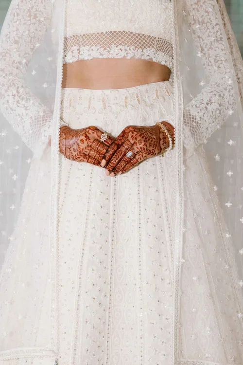 Close up of a brides hands covered in henna with diamond-rings and an embroidered-lace dress and long veil