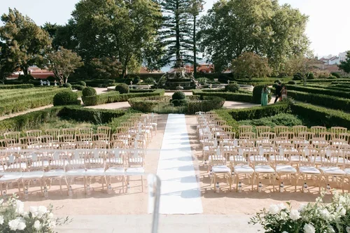 Outdoor wedding ceremony chairs set in rows along a white aisle through formal box hedge parterre gardens with tall trees behind at a grand Lisbon wedding venue