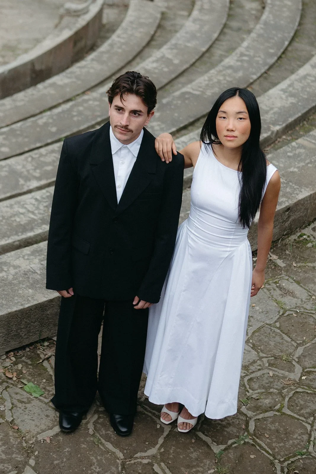 Bride in a white asymmetric midi dress and groom in a black suit standing on ancient stone steps photographed from above in Malaga