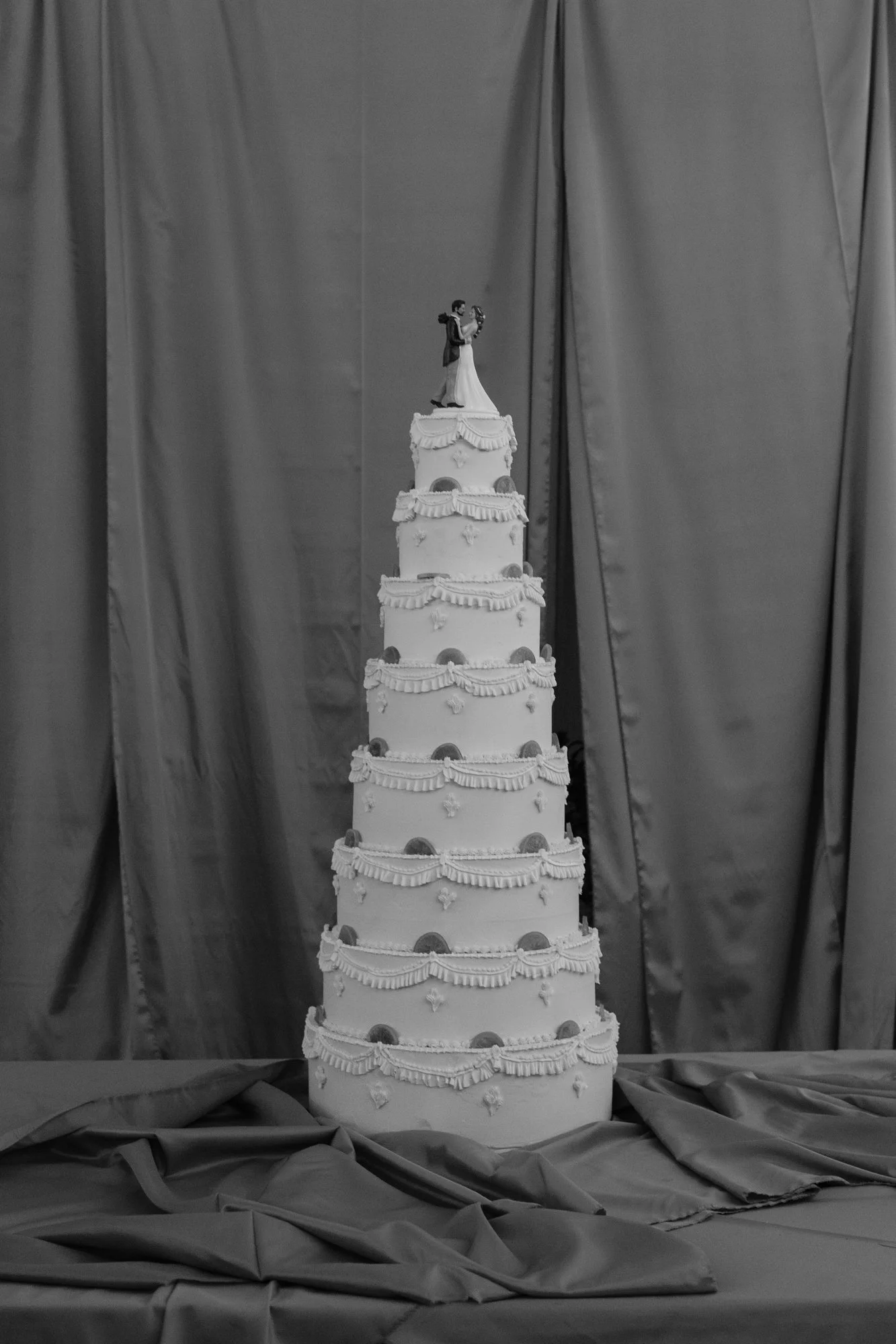 Tall eight-tier ornate white wedding cake with bride and groom topper against a draped grey backdrop