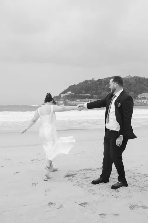 Couple dancing playfully on a beach in Marbella, groom spinning bride in white dress with waves and hillside town behind