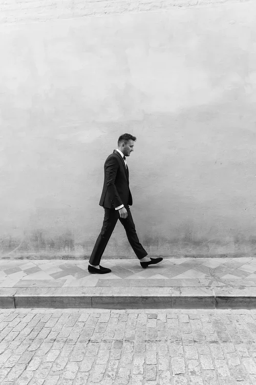 Groom in black tuxedo walking confidently along a wide whitewashed Andalusian street with cobblestones and geometric tiled pavement