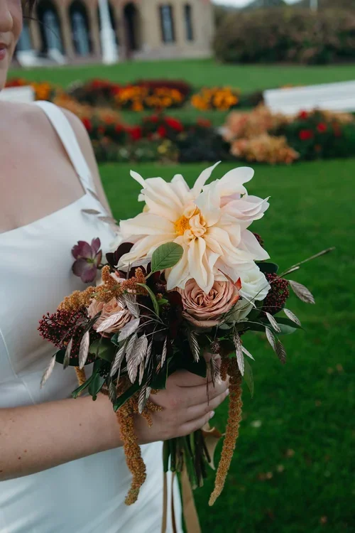 Bride holding a large autumnal wedding bouquet of café au lait roses, blush dahlias, dark burgundy foliage and hanging amaranthus against a white dress and garden backdrop