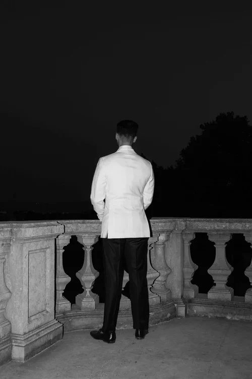 Groom in white dinner jacket standing with his back to camera looking out over a dark garden from a stone balustrade balcony at night