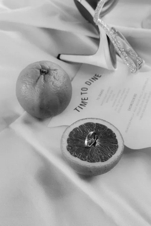 Styled flat lay of wedding rings resting in a halved grapefruit beside bridal heels and a menu card reading Time To Dine