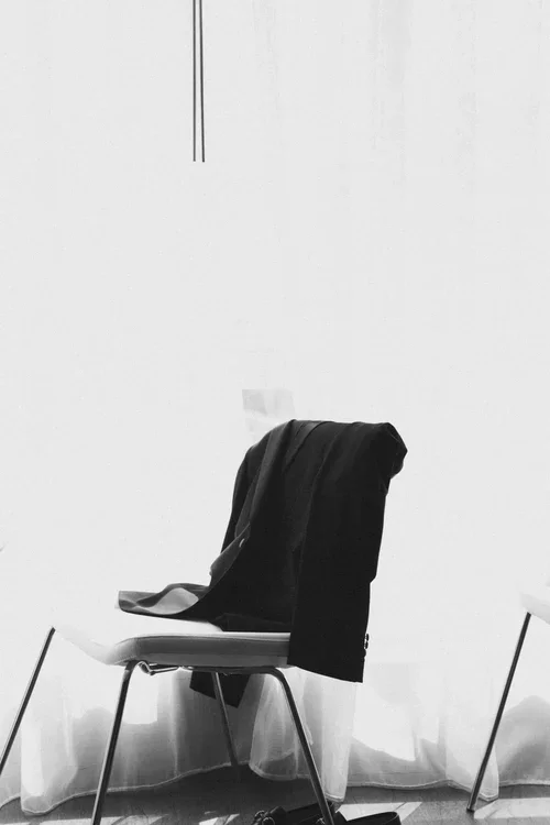 Black tuxedo jacket draped casually over a white chair against sheer white curtains in a Malaga hotel room on a wedding morning