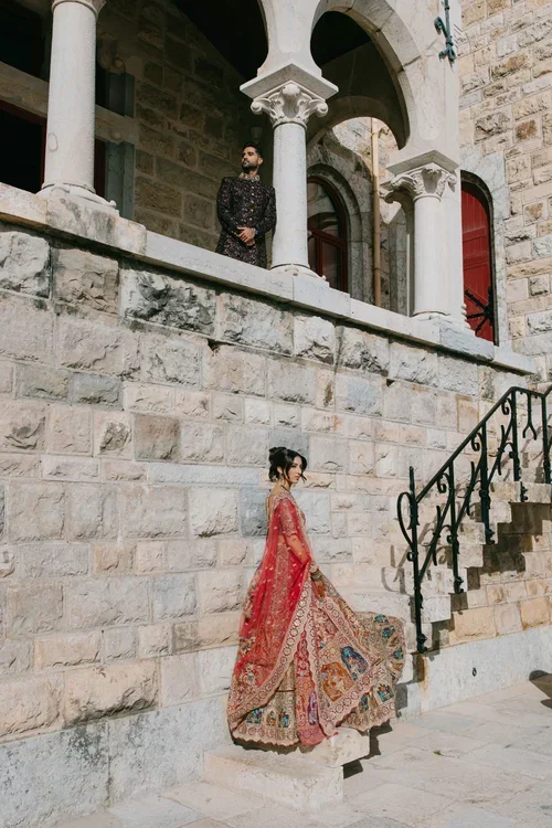 Bride in sweeping red and gold lehenga twirling on stone castle steps while groom watches from a balcony above at a Marbella castle wedding venue