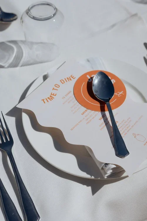 Stylish wedding place setting with wavy white plate, orange Time to Dine menu card and silver cutlery in Malaga