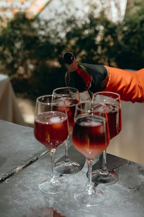 Waiter in red jacket pouring red wine sangria into multiple glasses lined up on a metal surface at an outdoor Malaga wedding reception