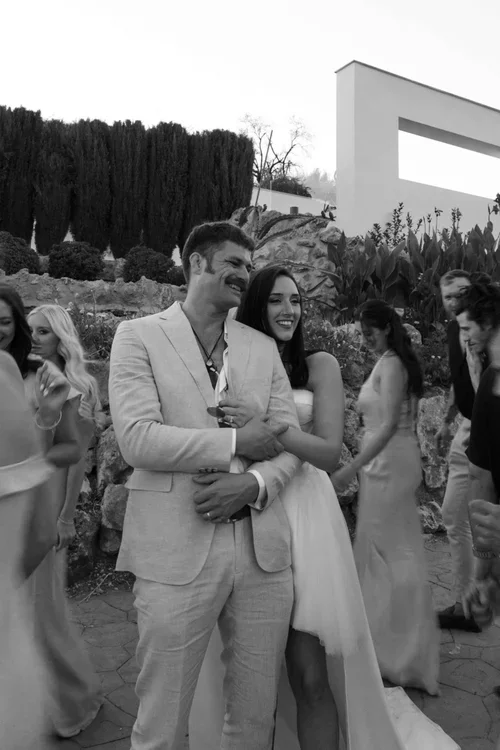 Bride in white strapless dress and groom in beige linen suit laughing together surrounded by guests at an outdoor wedding reception with rocky garden terracing behind them