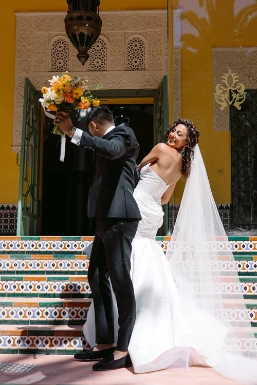 Smiling bride in backless mermaid dress with cathedral veil and groom in black tuxedo posing joyfully on ornate Moorish tiled steps at a Seville wedding venue
