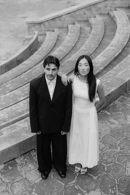 Bride in white asymmetric dress and groom in black suit standing together on ancient curved stone steps in Malaga