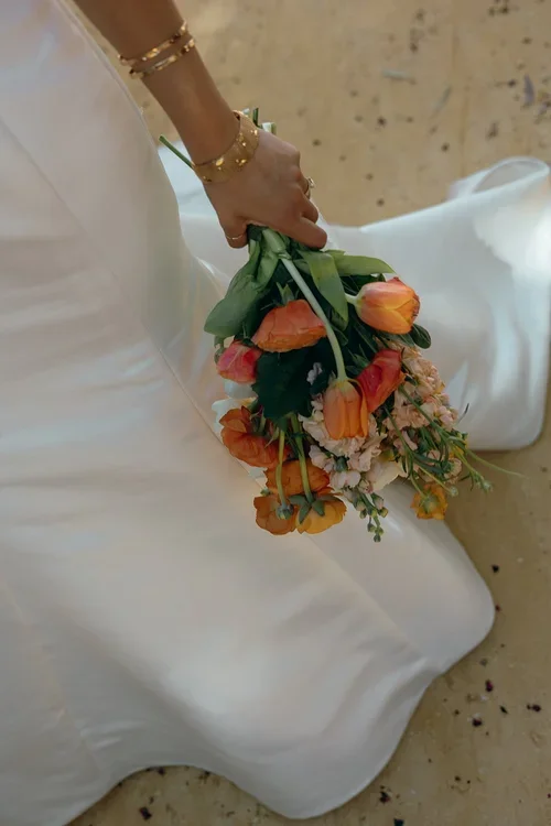 Close-up of bride's hand with gold bangles and rings holding a loose orange tulip and poppy bouquet against a white silk wedding dress