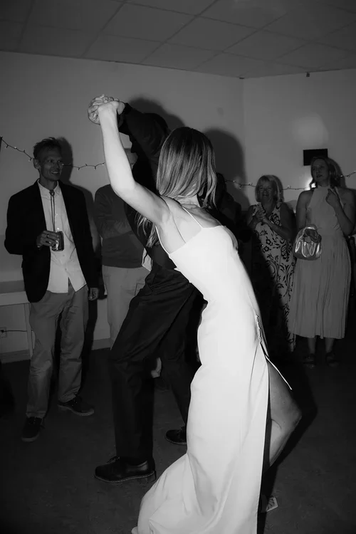 Bride in silk slip dress and groom dancing energetically together at an intimate wedding evening reception while guests watch and smile