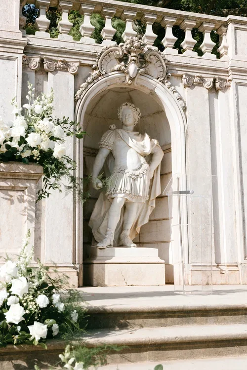 Classical marble statue in a carved stone niche flanked by lush white floral arrangements at an outdoor wedding ceremony in the Malaga hills
