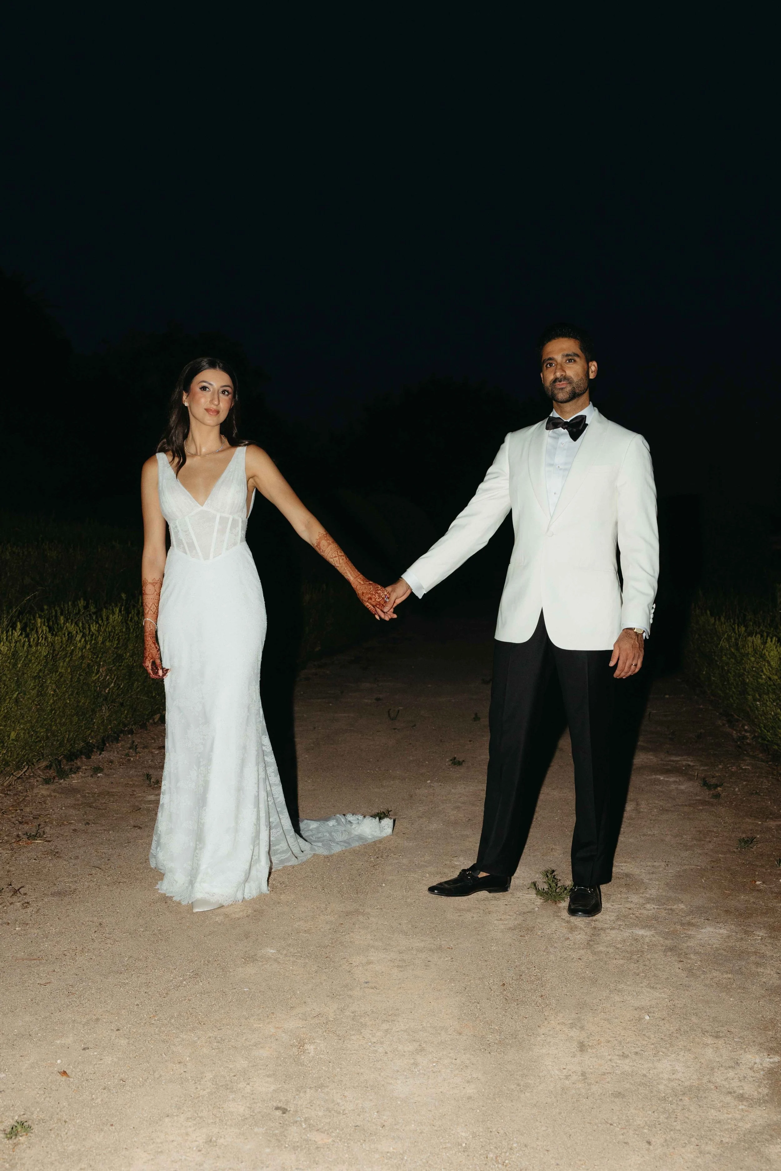 Bride in fitted lace wedding dress with henna arms and groom in white dinner jacket holding hands at night in Marbella