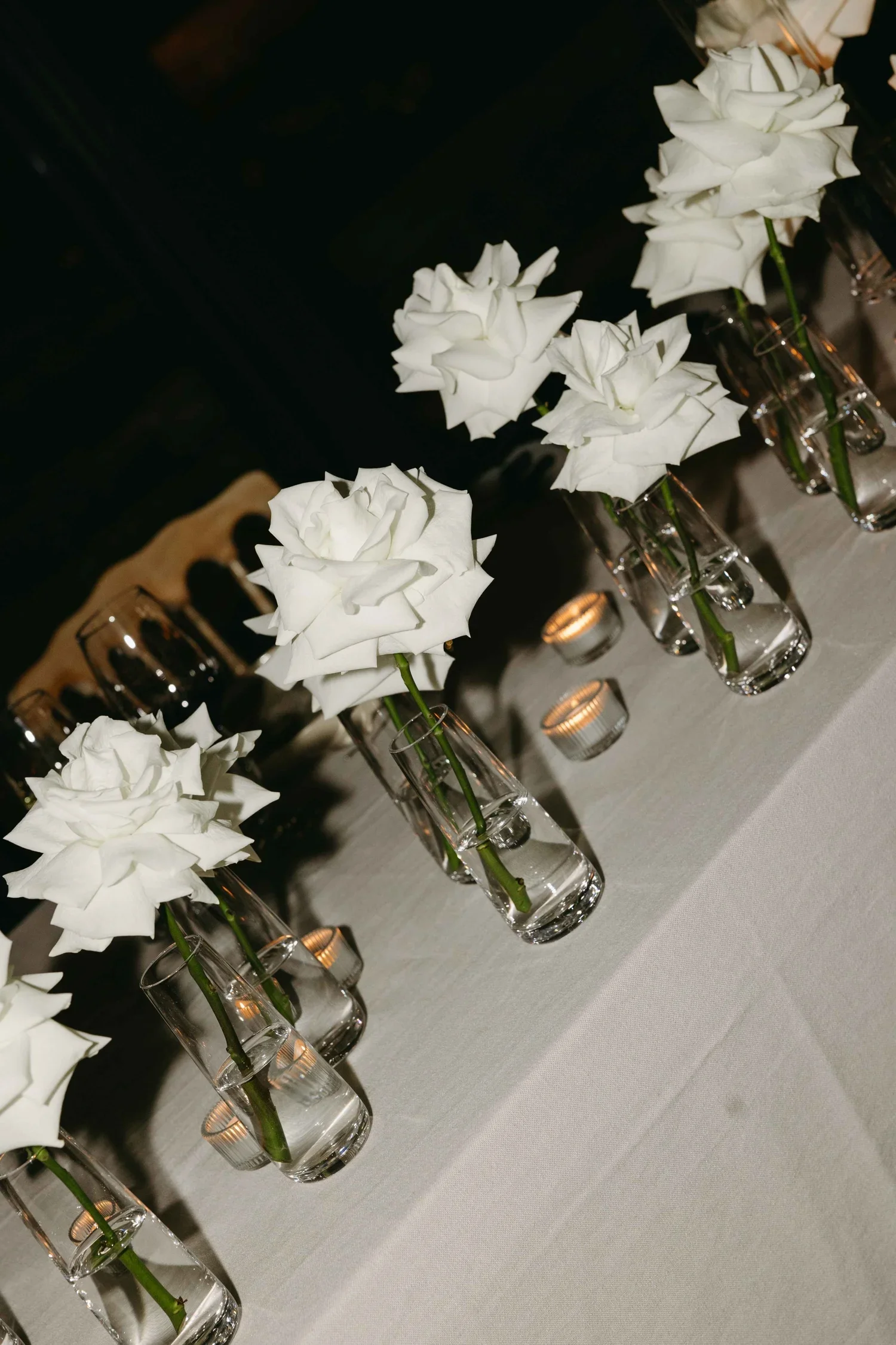 Single white roses in glass bud vases with tea light candles along a wedding reception table