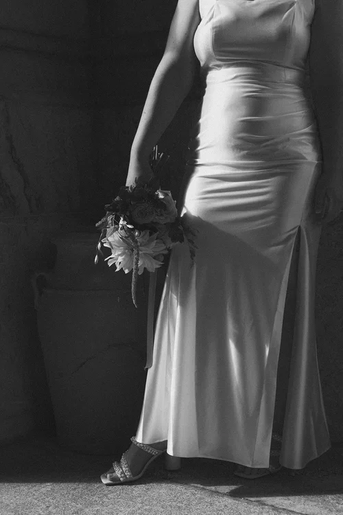 Close crop of bride in bias-cut silk wedding dress holding a wildflower bouquet against a dark stone wall