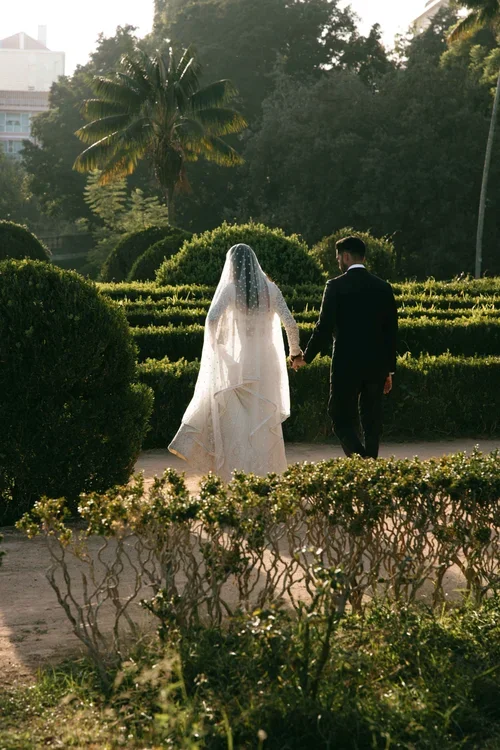  Bride in long veil and groom walking through tropical formal gardens at golden hour in Malaga 