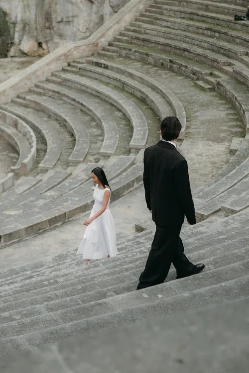 Bride in white midi dress and groom in black suit walking across the ancient Roman amphitheatre steps in Malaga