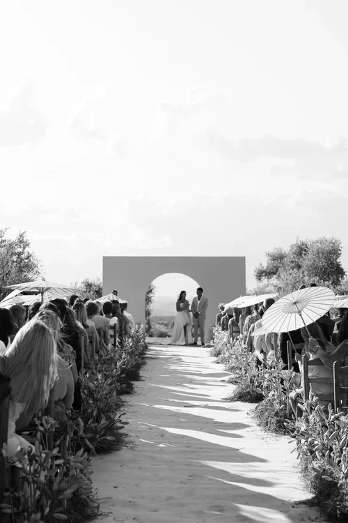 Wide shot of outdoor wedding ceremony in the Malaga hills with couple at a modern arch altar, guests seated along a sandy aisle shaded by white parasol umbrellas