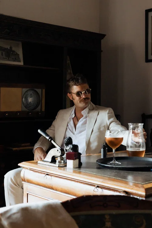 Groom in beige linen suit and sunglasses relaxing at an ornate antique wooden desk with a drink