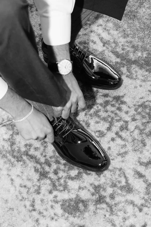 Close-up of groom lacing polished patent black Oxford shoes with a luxury watch on his wrist