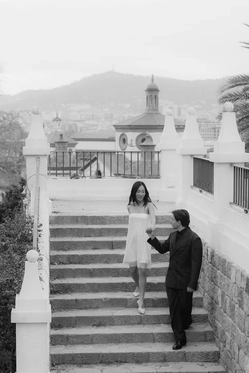 Bride in short white dress and groom in black suit walking down white stone steps with the Barcelona skyline and Tibidabo hill visible behind them