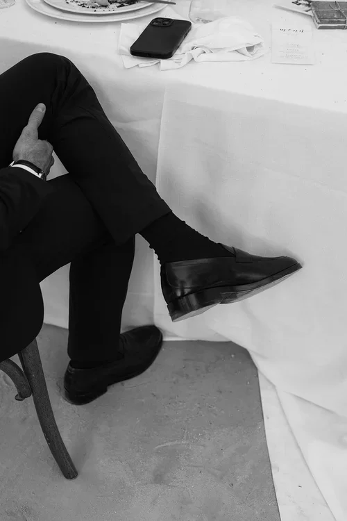 A guest with crossed legs wearing black suit trousers and designer loafers sitting at a table with a long draping white table cloth