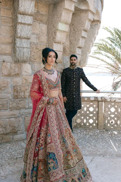 Bride in red and gold lehenga with sapphire jewellery and groom in dark embroidered sherwani posing at a coastal stone wedding venue in Marbella with the sea in the background