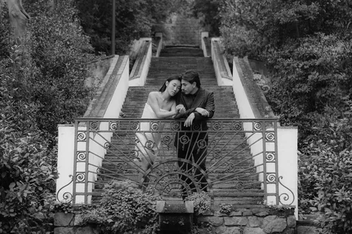 Bride and groom leaning intimately together against an ornate wrought iron garden railing surrounded by lush green climbing plants and a long staircase behind them
