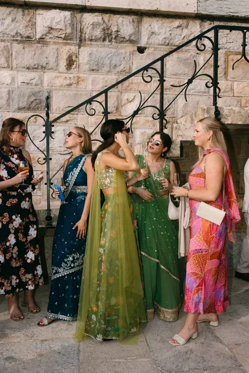Wedding guests in colourful Indian sarees and Western dresses laughing at a drinks reception against a stone wall in Marbella