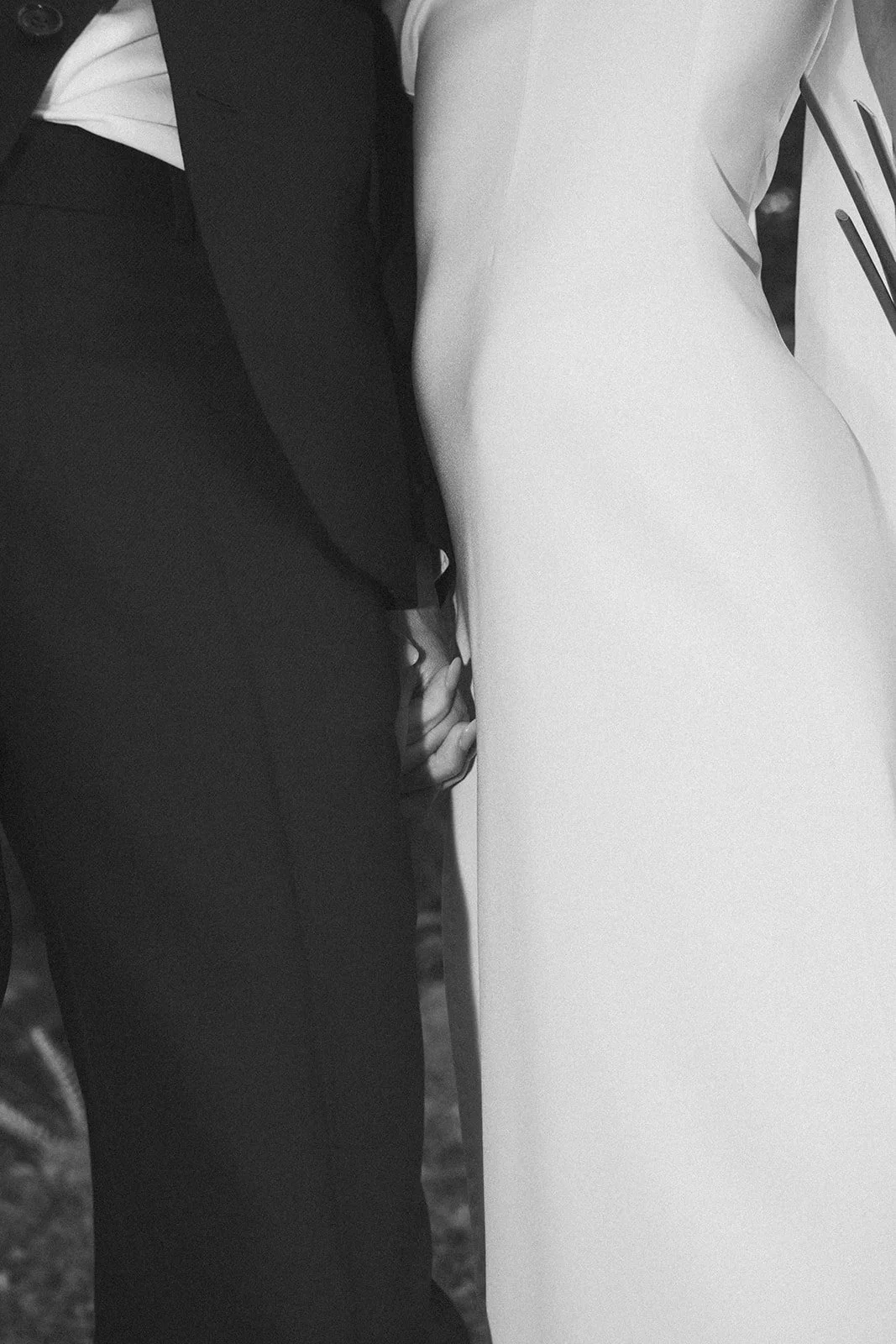 Intimate close-up of bride and groom holding hands, white dress beside dark suit