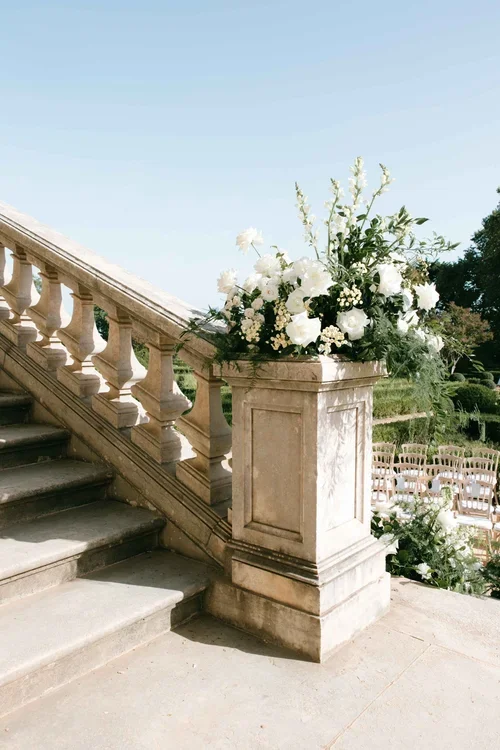 Carved stone balustrade staircase with a large white floral arrangement of roses and peonies on a stone plinth at an outdoor Seville wedding ceremony