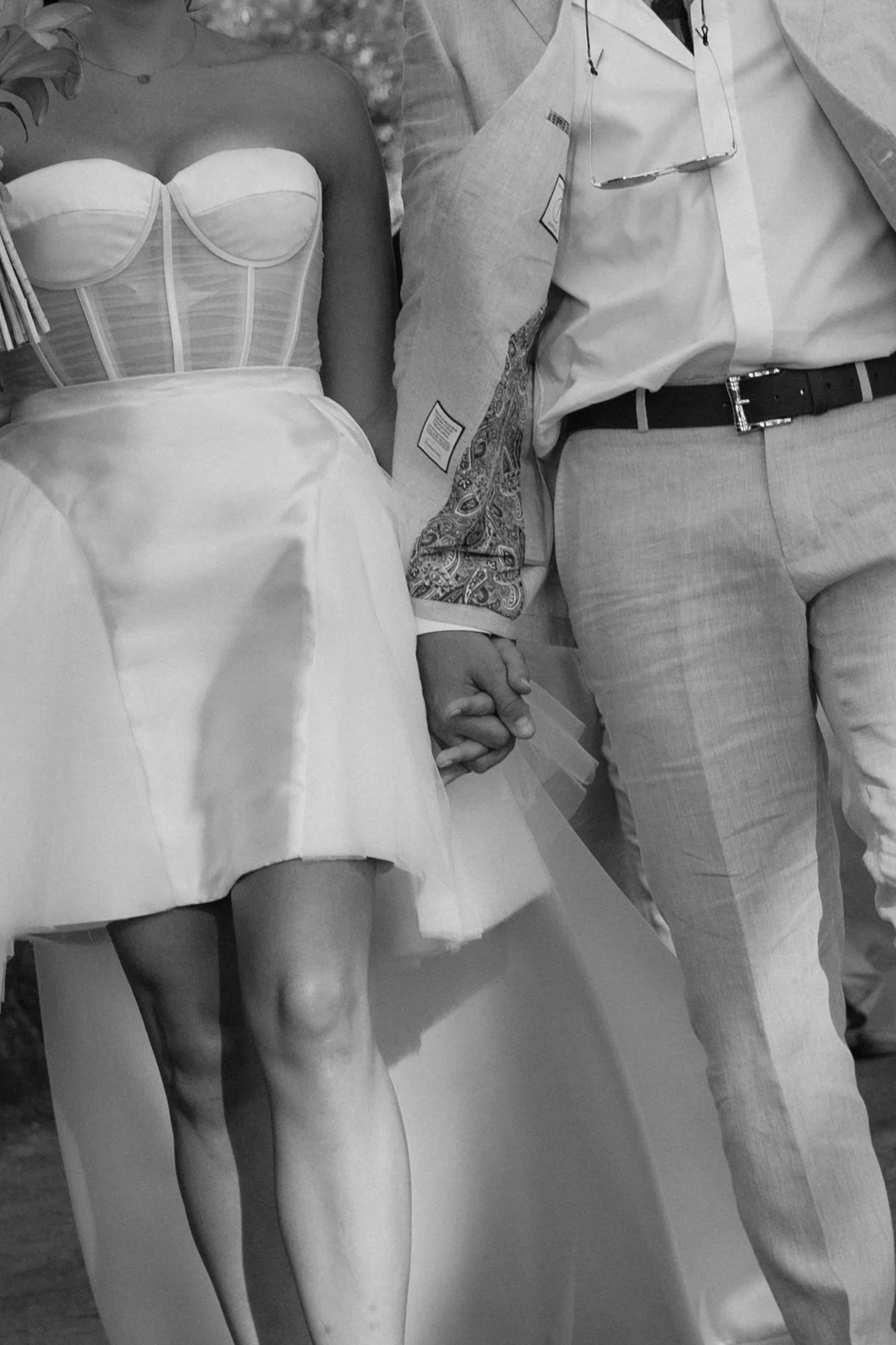 Close-up of bride in strapless corset mini dress and groom in linen suit holding hands at their wedding