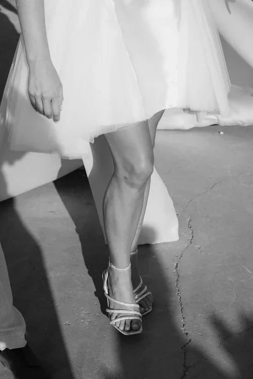 Close-up of bride wearing embellished strappy heeled sandals beneath a floaty white dress mid-stride