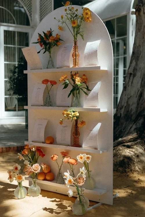 Styled white arched shelving unit seating plan display with orange ranunculus alstroemeria and daffodil bud vases and handwritten table plan cards at a Marbella wedding venue