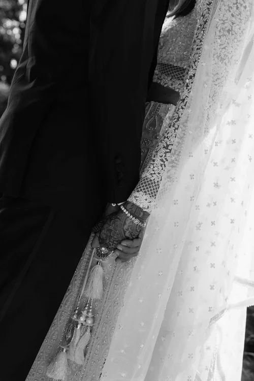 Close-up of bride and groom holding hands with henna decorated wrist bracelet and tassel kalire charm visible against embroidered lace sleeve and star-embroidered veil, black and white wedding detail photography Marbella