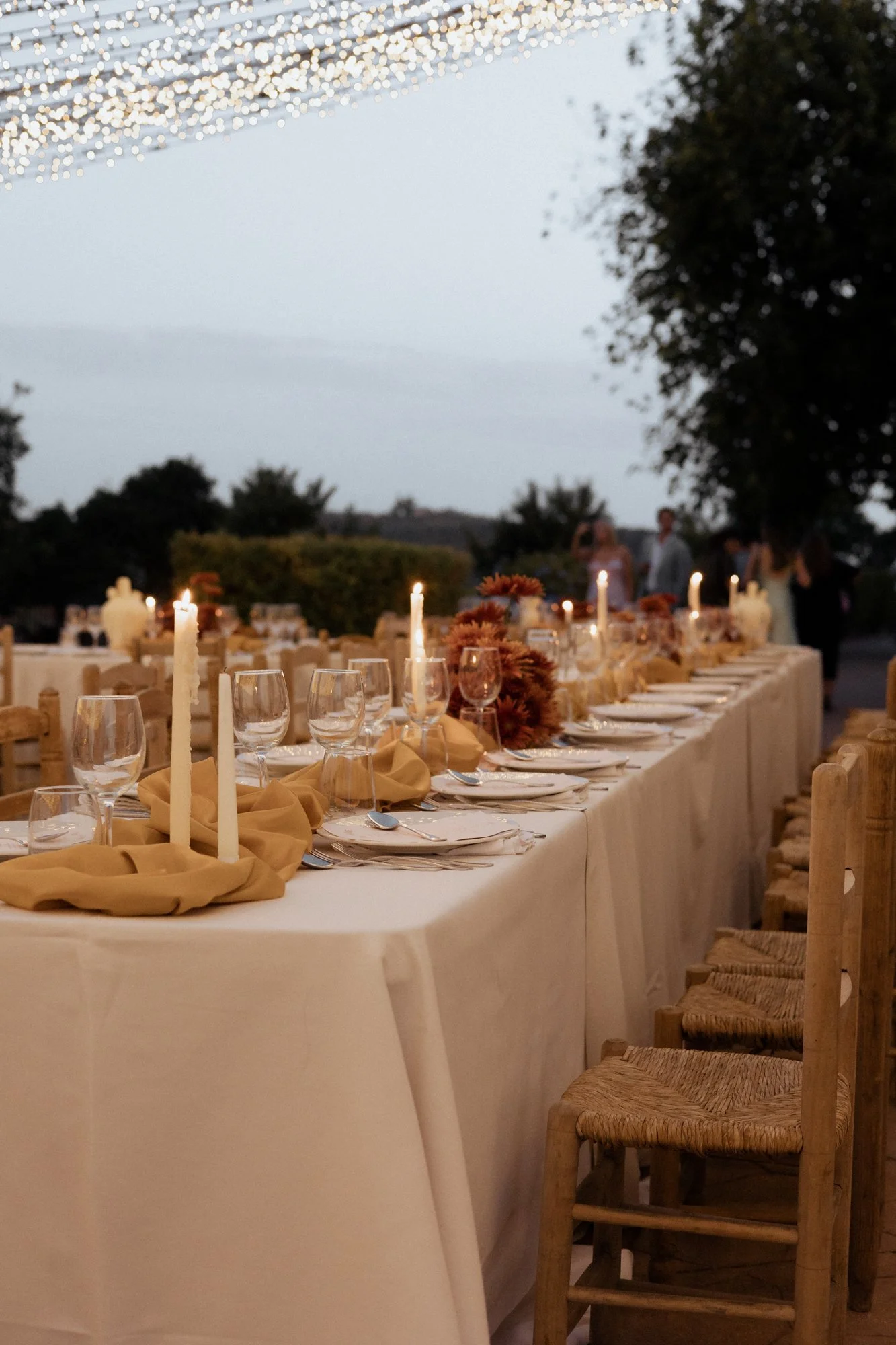 Outdoor wedding reception table set for dinner at dusk with candles, mustard napkins and fairy lights in Marbella