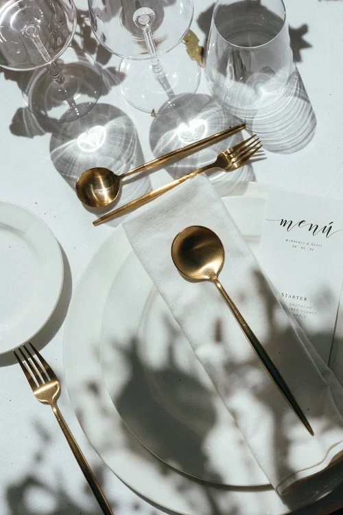 Elegant wedding place setting with white plate, gold cutlery, crystal glassware and printed menu card in bright sunlight