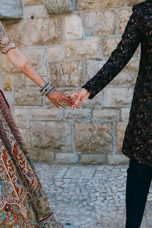 Close-up of bride's henna-decorated hands with blue bangles touching groom's hand against a stone wall