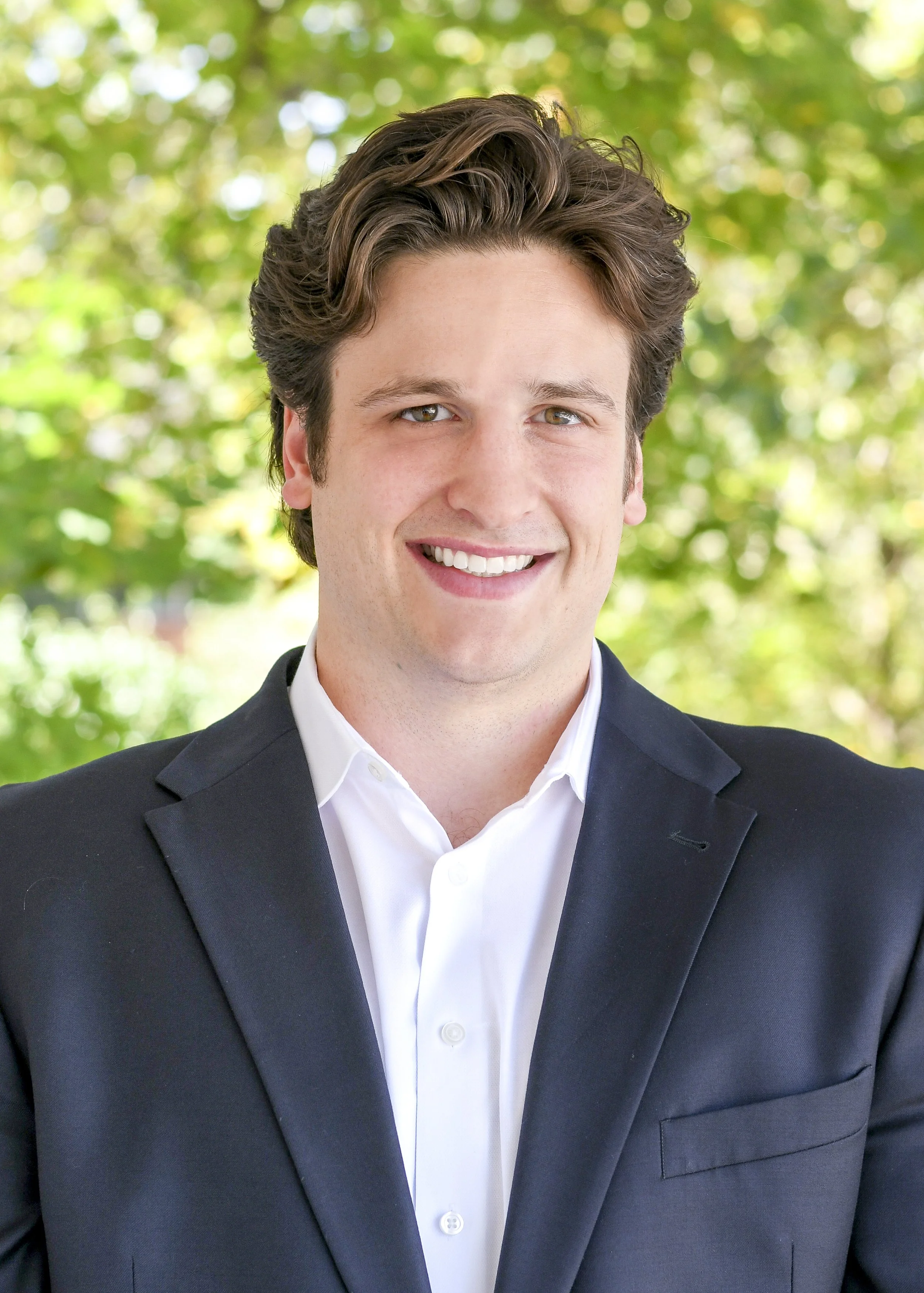 A smiling young man with dark brown hair in a white dress shirt and black blazer, standing outdoors with green trees in the background.