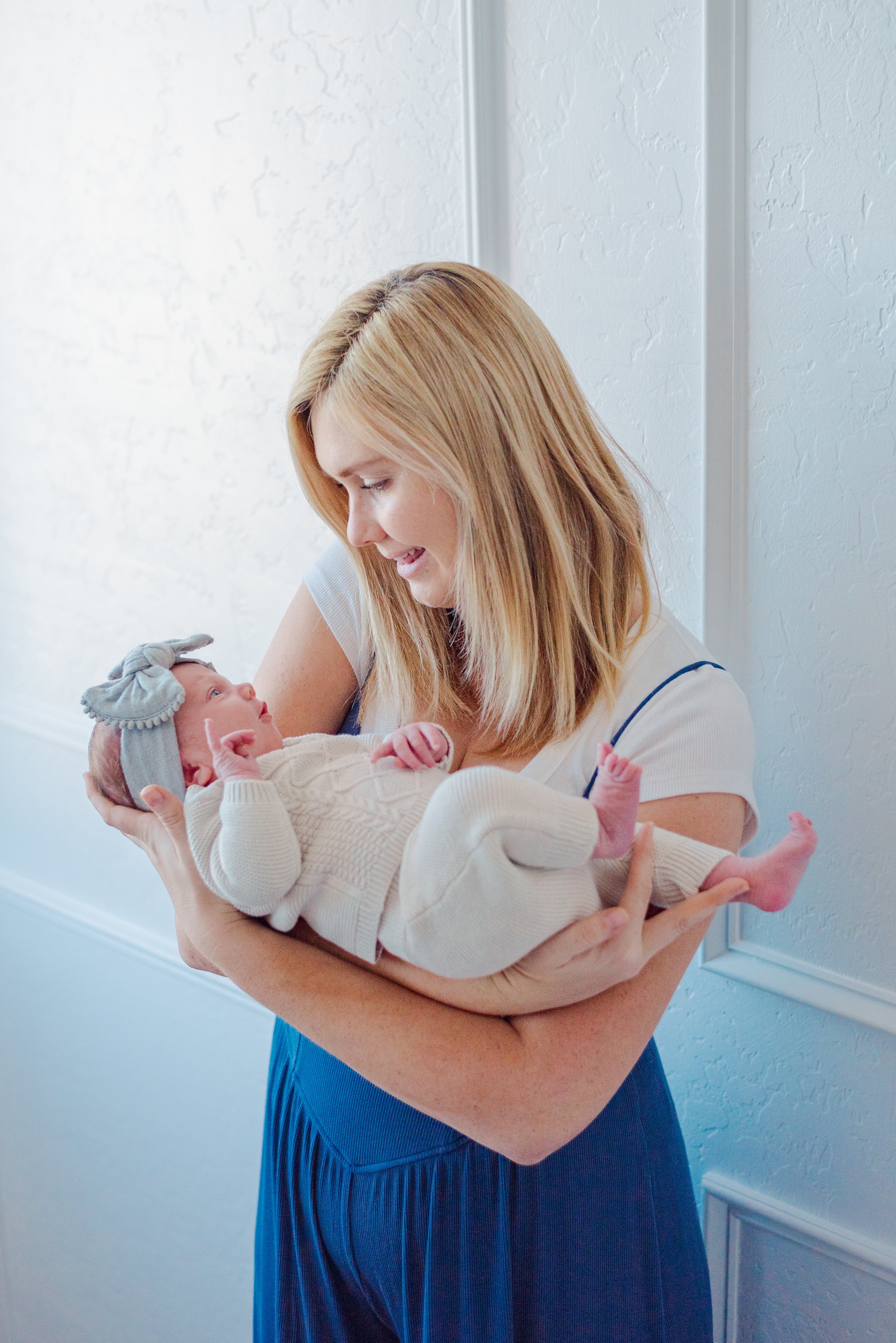 A mother holds her baby girl in her arms against a white wall background.