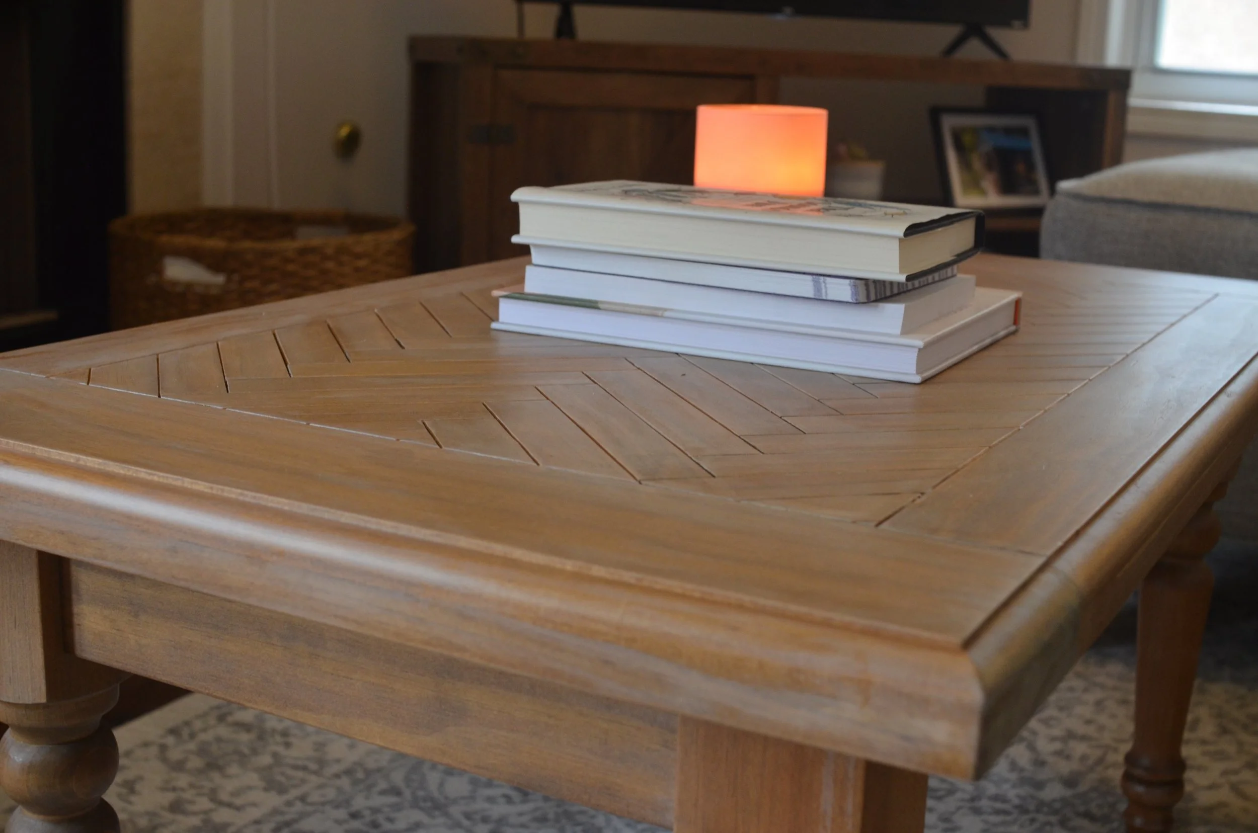 Stack of four books on a wooden table in a living room, with a lit pink lamp behind them.