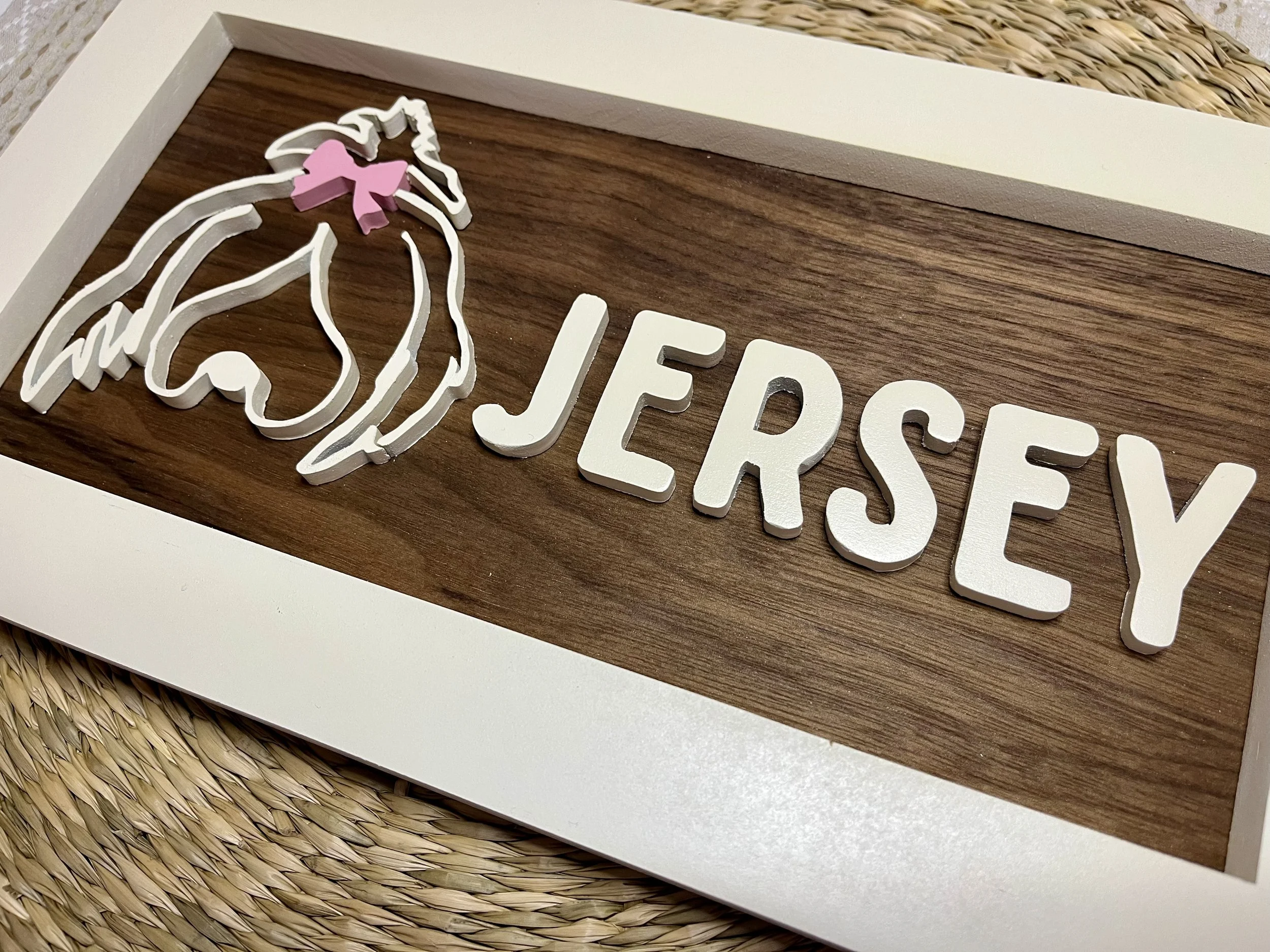 Decorative wall art with the word 'JERSEY' in white 3D letters and a white outline of a horse's head with a pink bow, mounted on a wooden background within a white frame.